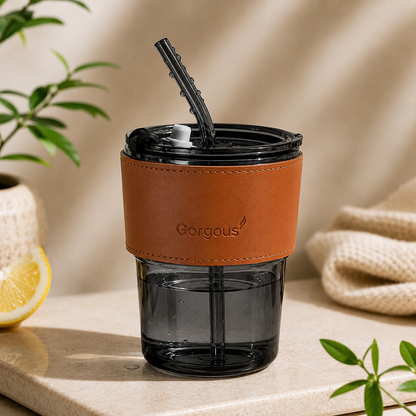 Black aesthetic glass tumbler with reusable straw and brown leather sleeve on neutral background