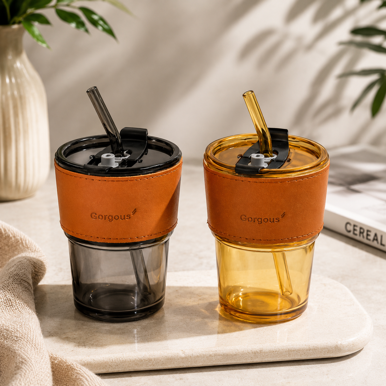 Gold and black aesthetic glass coffee tumblers with reusable straws and brown leather sleeves on neutral background