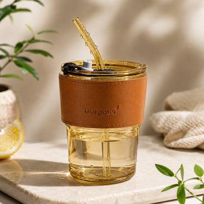 Gold aesthetic glass tumbler with water, reusable straw and brown leather sleeve on beige background